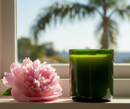 Chez Shea luxury candle in a green vessel beside a pink peony on a sun-lit windowsill with palm trees in the background