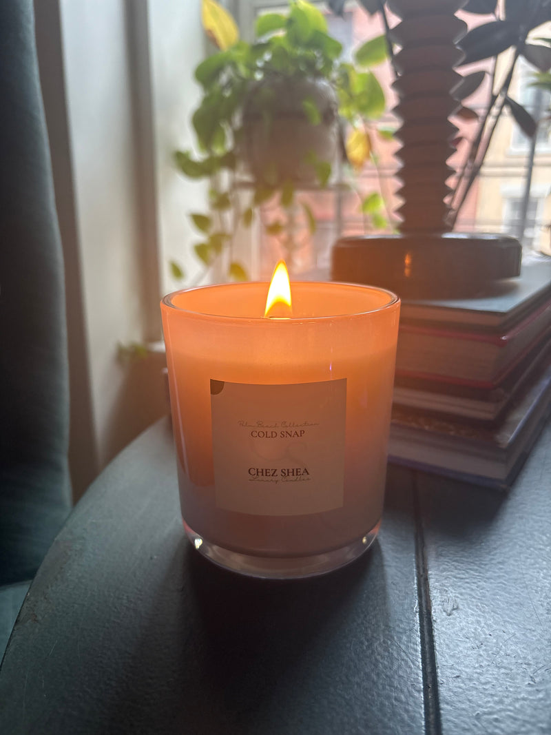 Candle with a visible brand label on a surface near a window with plants.