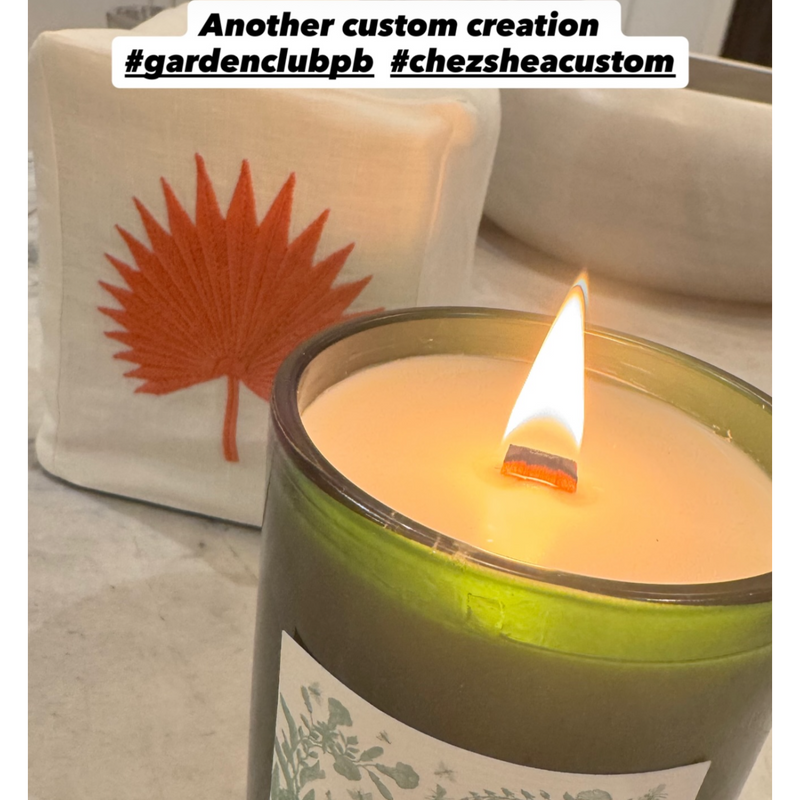 Candle with a label on a surface, with a bag featuring a leaf design in the background.